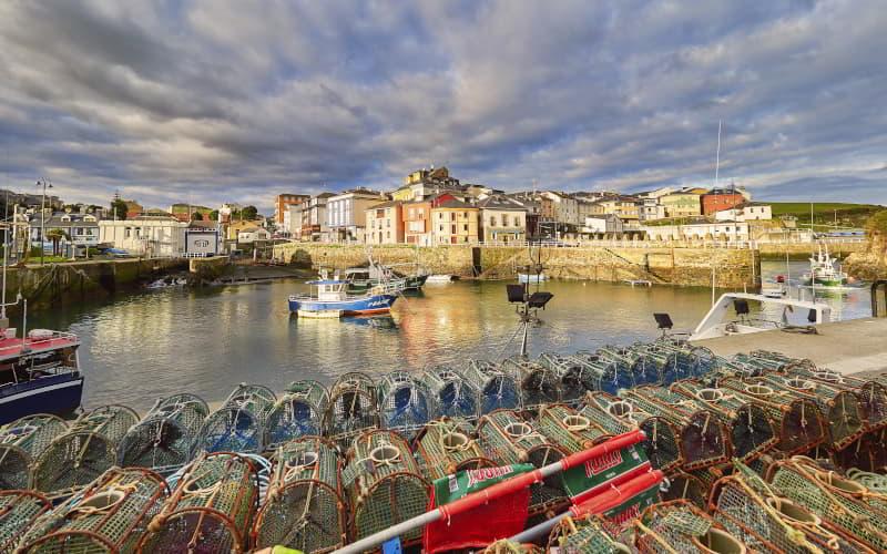 Vista del puerto de Puerto de Vega con embarcaciones pesqueras atracadas y nasas de marisco apiladas en primer plano. Al fondo se observan las fachadas de las casas del pueblo junto a la dársena, bajo un cielo parcialmente nublado.