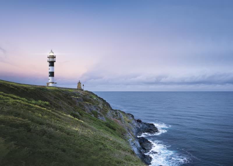 Faro de San Agustín, torre blanca y negra de franjas horizontales, situado sobre un acantilado verde frente al mar Cantábrico. El faro emite luz al atardecer, con el cielo teñido de tonos rosados y azulados sobre el horizonte.