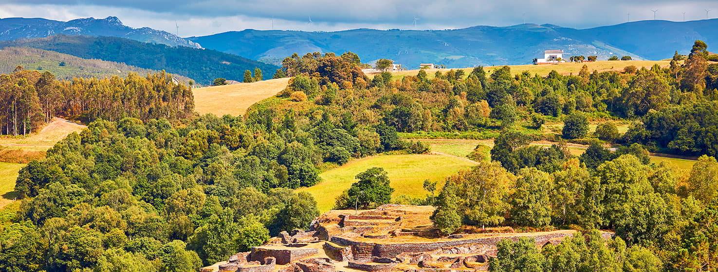 Panoramic view of Castro de Coaña, with archaeological remains of circular structures and walls in the foreground, surrounded by green meadows and groves. In the background are hills and mountains under a clear sky.
