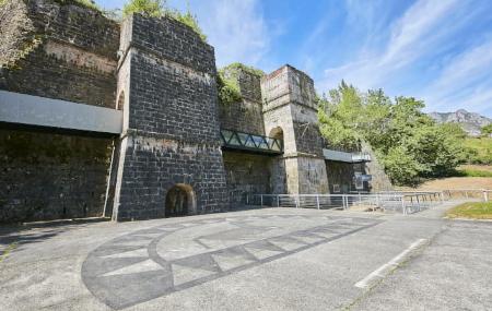 Estructuras de piedra de los antiguos altos hornos de fundición en Quirós, con muros elevados, arcos y pasarelas metálicas añadidas para su musealización. En primer plano se observa una amplia explanada con motivos geométricos en el pavimento, rodeada de vegetación y montañas al fondo bajo cielo despejado.