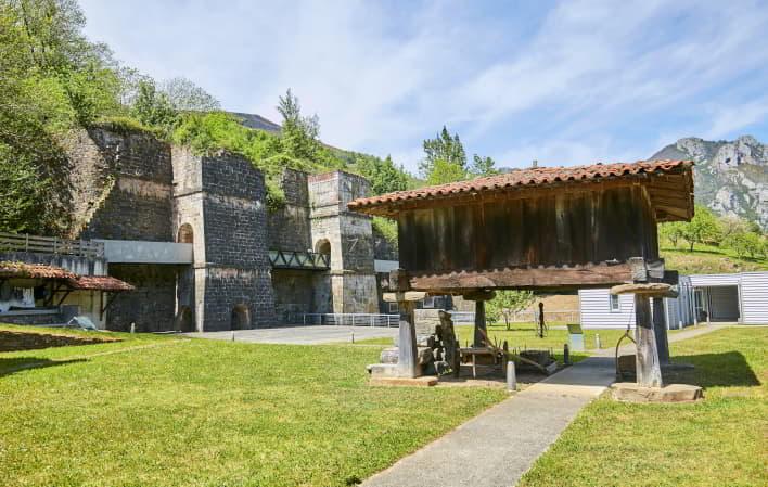 Hórreo tradicional asturiano en primer plano dentro del recinto del Museo Etnográfico de Quirós, con los antiguos altos hornos de fundición al fondo y montañas rodeadas de vegetación bajo cielo despejado. Museo Etnográfico de Quirós