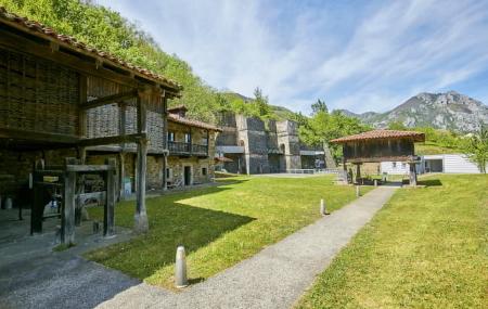 Conjunto del Museo Etnográfico de Quirós, donde se integran hórreos y edificaciones tradicionales de piedra junto a las estructuras de los antiguos altos hornos de fundición. El entorno está rodeado de praderas verdes y montañas, bajo un cielo despejado con nubes finas.