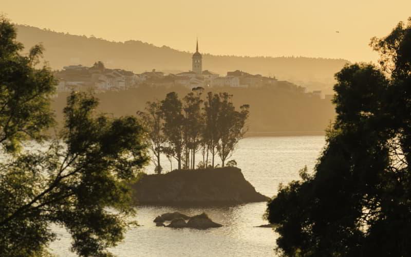 Coucher de soleil dans l'estuaire de l'Eo, avec une petite île boisée au premier plan et la silhouette de la ville de Castropol et de son église à l'arrière-plan, enveloppée d'une lumière dorée.