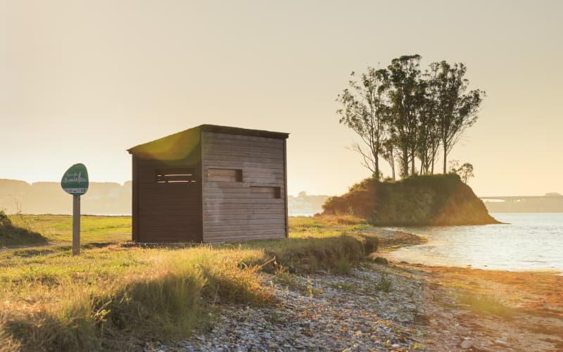 Observatoire d'oiseaux en bois près de l'estuaire de l'Eo, sur la plage de Salías (Castropol), avec une petite île boisée en arrière-plan et la lumière chaude du coucher de soleil.