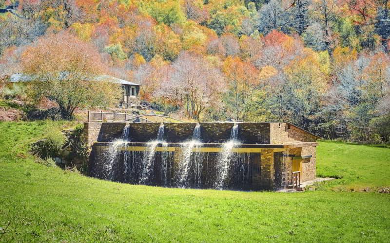 La Ferrería de Santa Eufemia à Villanueva de Oscos, un bâtiment en pierre avec de l'eau en cascade le long de sa façade, entouré de prairies et de bois aux couleurs automnales.
