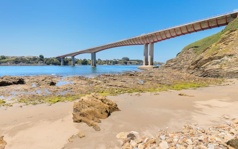 Vue du pont sur la rivière Eo depuis la plage de San Román à Castropol, avec au premier plan le sable, les rochers et les zones de marée basse, sous un ciel dégagé.