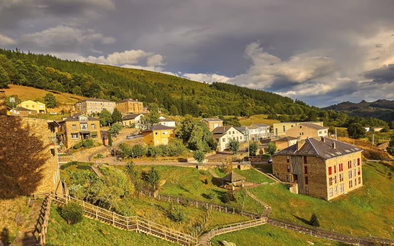 Vue du centre de Santa Eulalia de Oscos avec des maisons en pierre et des toits en ardoise sur des pentes vertes, entourées de forêts et de montagnes dans la lumière chaude de l'après-midi.