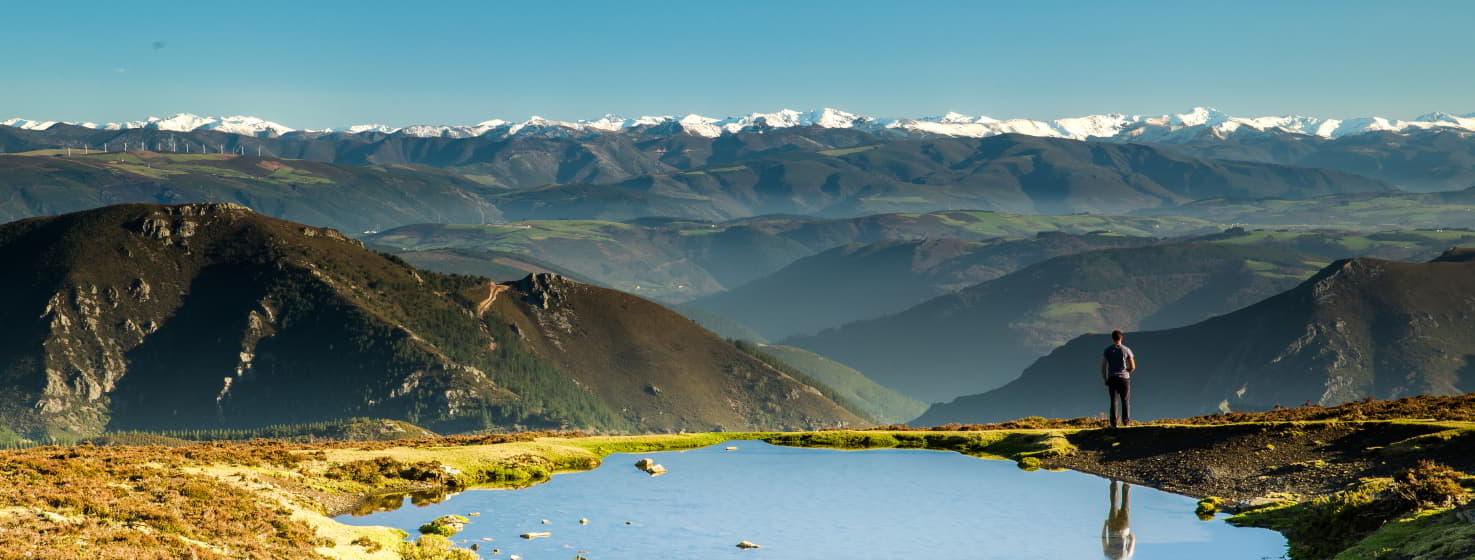 Vue panoramique de Villanueva de Oscos montrant un paysage montagneux de La Bobia avec un ciel clair. Au premier plan, un bassin d'eau reflète l'environnement, tandis qu'un homme contemple la vue. À l'arrière-plan, une chaîne de montagnes enneigées s'étend sur un horizon de vallées et de collines, créant un beau contraste entre le vert et le blanc.