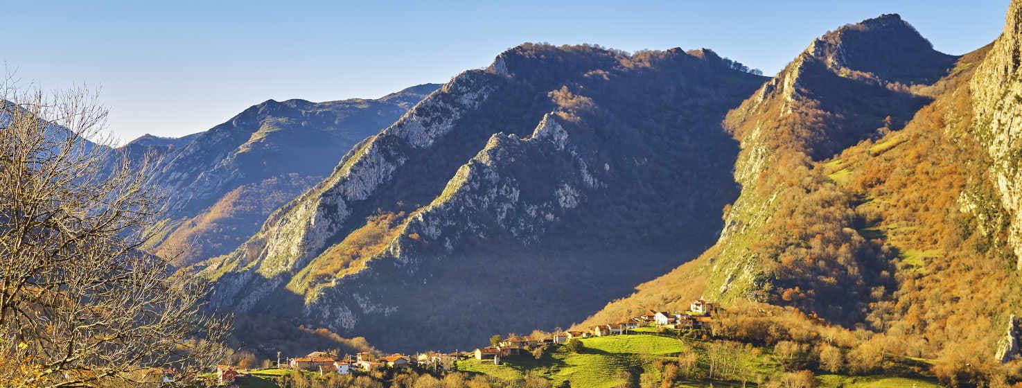 Impressionante paisagem de montanha com a vista da pequena aldeia de Pen, Amieva, no centro da fotografia. Rodeada de prados e árvores, neste caso com cores de outono, predominando os tons laranja e ocre das folhas. Ao fundo, montanhas espectaculares erguem-se em direção ao céu com formas caprichosas, umas mais nítidas e outras mais arredondadas.