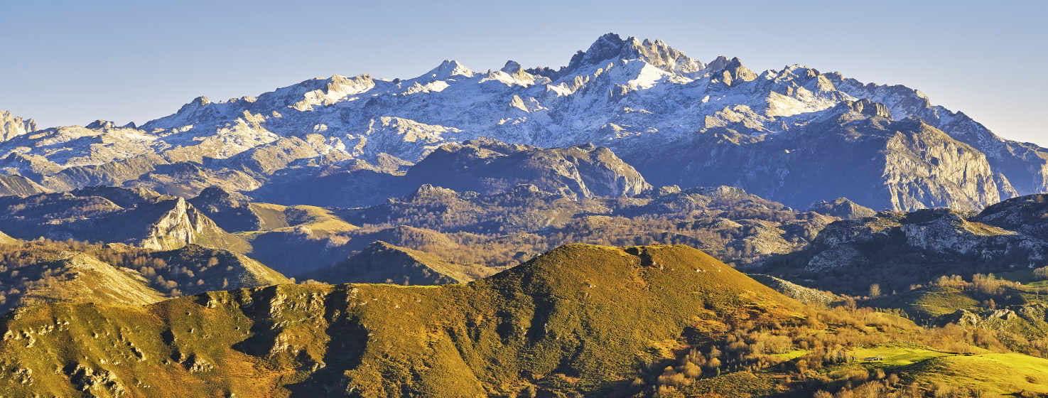 Vista do maciço ocidental dos Picos da Europa, com neve nos picos e montanhas um pouco mais baixas em primeiro plano, diferentes tons de verde dos prados, combinados com o cinzento da rocha e o branco da neve. O céu azul confere grande luminosidade à paisagem.