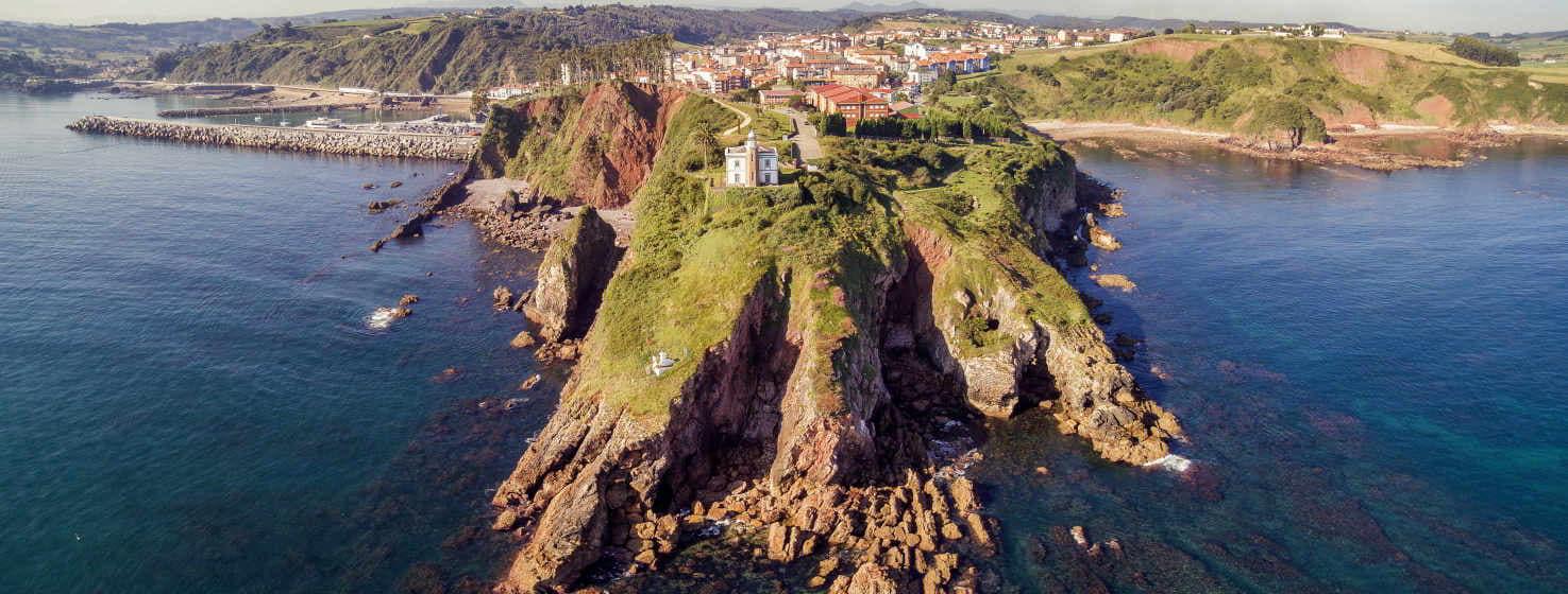 On voit le cap s'enfoncer dans la mer avec d'importantes falaises, au sommet le phare, blanc, sur une prairie verte, en arrière plan on voit le village de candas avec des plages à droite et le port à gauche de l'image. la mer est bleue et calme.