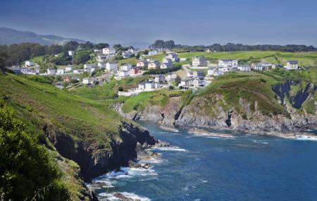 Vista de Coaña en lo alto del acantilado, no muy alto. Pequeño pueblo de casas blancas y tejados de pizarra. A sus pies el mar cantábrico que se adentra como una pequeña lengua hacia el interior donde estaría su pequeño puerto. Al otro lado más acantilados de roca con praderías encima.