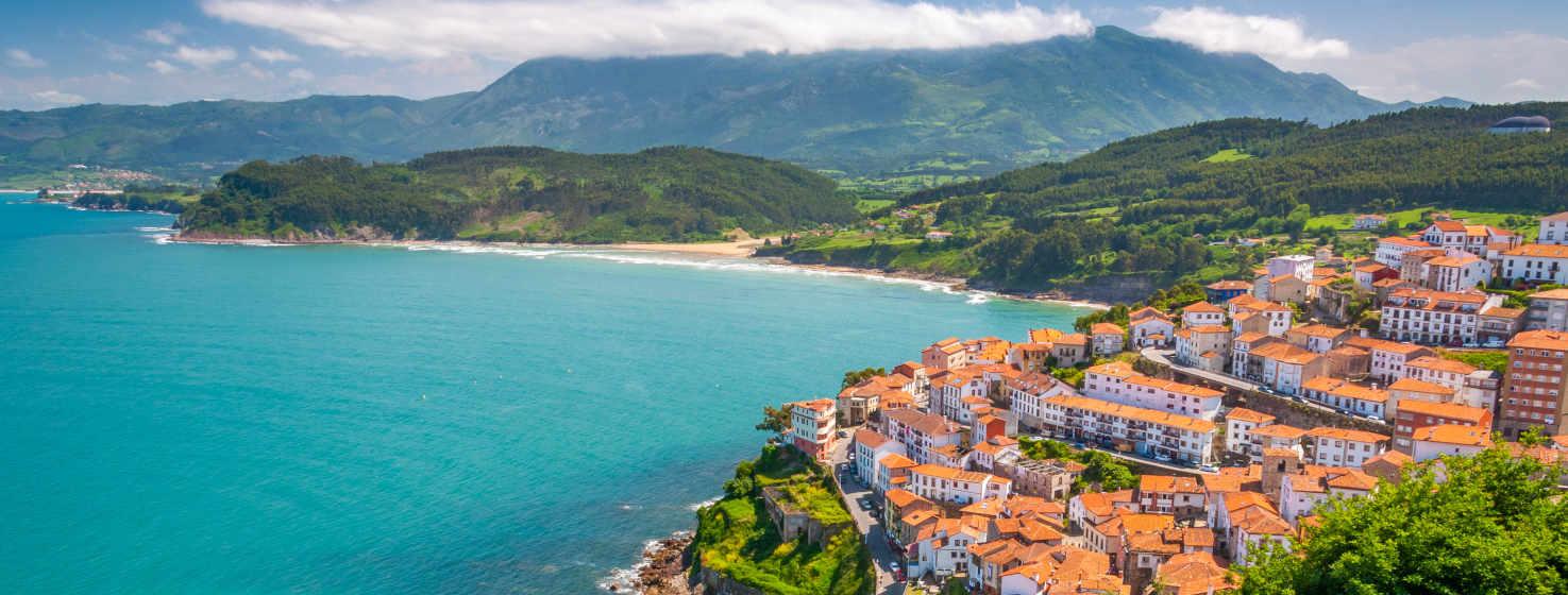 Bellissima vista di Lastres con le sue case bianche e rosse che formano un balcone sul mare. Il mare è trasparente, si può vedere lo sfondo vicino alla riva più vicina al villaggio. Dall'altra parte si vede la spiaggia di La Griega e sopra di essa morbide colline verdi con alberi. Dietro, la bluastra Sierra del Sueve con la sua appuntita cima Pienzu.