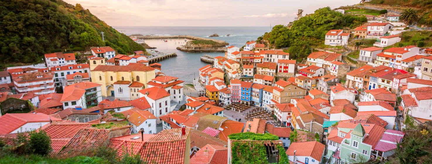 Image du village de Cudillero, au premier plan on peut voir un groupe de maisons aux toits rouges serrées les unes contre les autres formant un amphithéâtre surplombant la mer, sur les côtés au-dessus du village se trouvent de douces collines avec des arbres bas.  Sur la côte, on peut voir le port et l'horizon en arrière-plan.