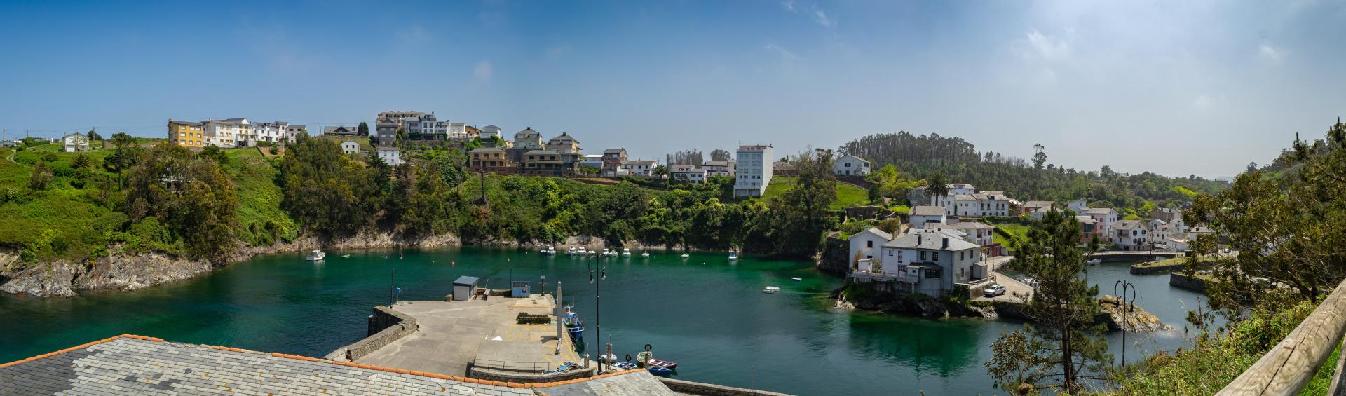 Imagem da aldeia piscatória de Viavélez, em primeiro plano o porto com o mar calmo e límpido, protegido das ondas. Podem ver-se pequenos barcos atracados. À direita da imagem, há um núcleo de pequenas casas brancas da aldeia, à esquerda, no cimo da colina, há mais casas, sob um céu azul.
