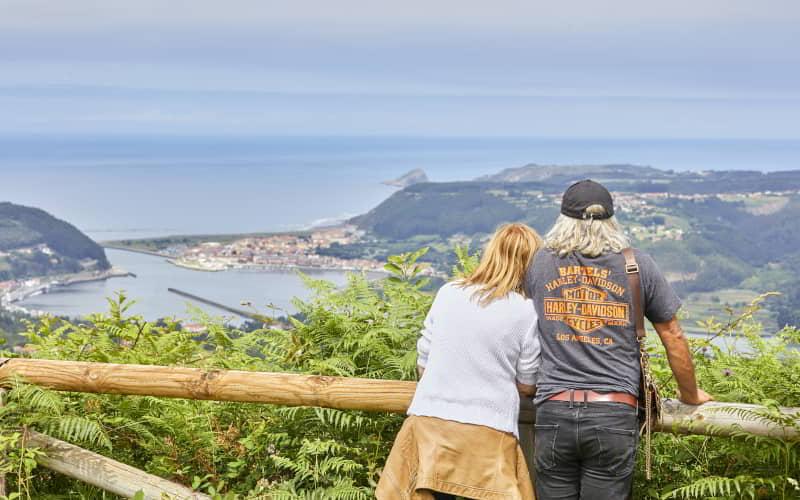 Dos personas observan desde un mirador de madera el paisaje costero donde el río Nalón desemboca en el mar. Al fondo se distinguen la villa de San Esteban y la costa cantábrica, con colinas verdes y cielo nublado.