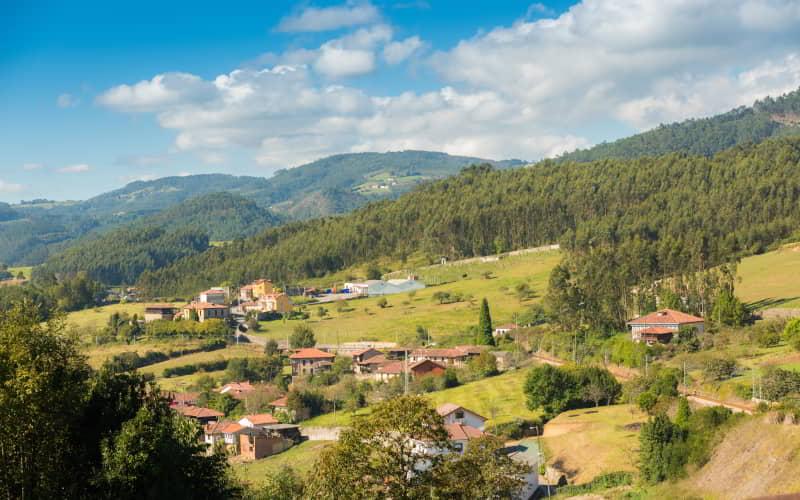 Vista panorámica de la localidad de Santianes, con casas rurales dispersas entre prados y colinas cubiertas de bosques. El paisaje muestra un entorno verde y montañoso bajo un cielo azul con nubes dispersas.