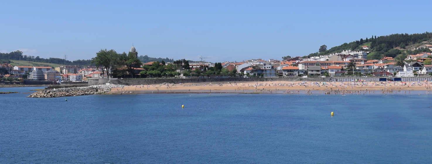 Vue de la mer sur la ville et la plage de Luanco. Les eaux très calmes, protégées par le port et la plage, vous pouvez voir une bande de sable doré avec des gens, derrière les maisons du village et les arbres, à gauche le Cabildo et une autre entrée de la mer au village qui atteint l'ancien quai.