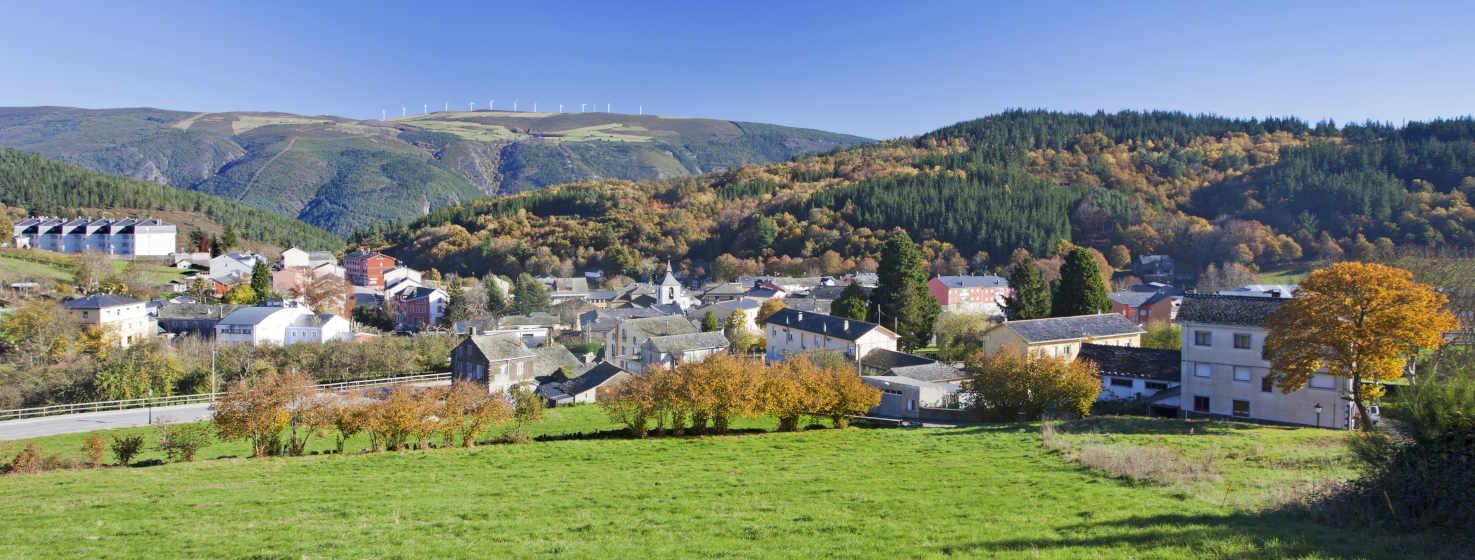 Vista de Grandas de Salime com uma paisagem montanhosa por detrás. A aldeia tem casas de pedra com telhados de ardósia e uma igreja com uma torre sineira. A paisagem é dominada por colinas cobertas de árvores e prados verdes. O céu está limpo e azul, com algumas nuvens brancas. A imagem transmite uma sensação de paz e tranquilidade.
