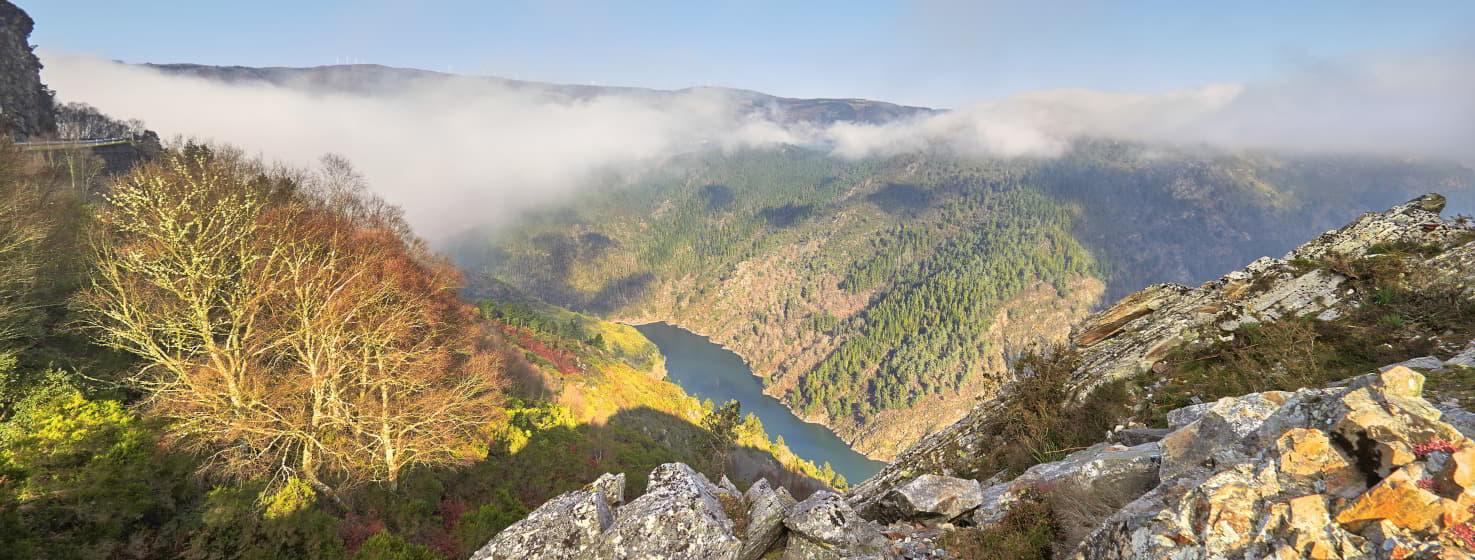 Fotografia da barragem de Dorias a partir do Mirador de los Buitres. Em primeiro plano, uma zona rochosa no cimo do miradouro, nos lados montanhas com árvores e arbustos em cores outonais, verdes, ocres, avermelhadas. Ao fundo, a barragem azul atravessa o vale.  Por detrás, mais montanhas a escovar o céu azul com uma ligeira névoa a subir do vale.