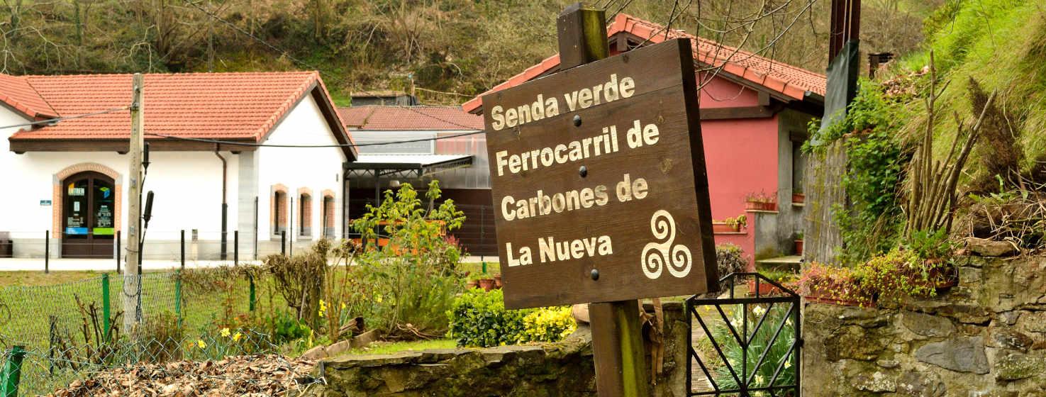 Imagen del comienzo de una senda verde, en primer plano un cartel de madera con el nombre de la senda verde y un trisquel adornando. Detrás el edificio blanco de la antigua estación de ferrocarril de La Nueva, ahora Eocumuseo de Samuño. Zona verde alrededor y otra casa roja a la derecha.