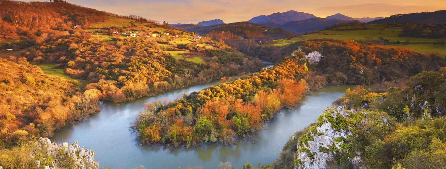 Imagem colorida dos Meandros do Nora. Do verde ao ocre, ao castanho, ao laranja, são as árvores que rodeiam o rio Nora. Paisagem totalmente outonal. No centro vemos um dos seus meandros, rio onde se reflectem as árvores. Ao fundo, podemos ver algumas montanhas azuis.