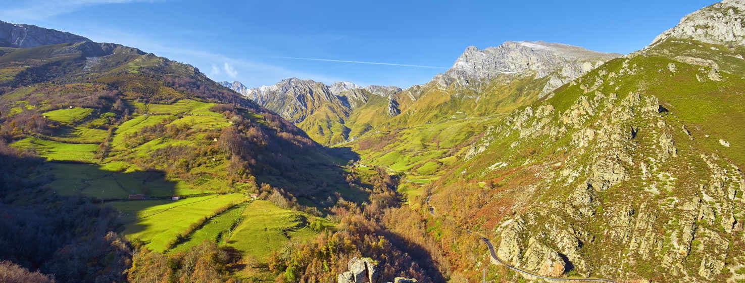 Photo d'un paysage de Lena. Des montagnes de différents verts avec des prairies délimitées et des arbres plus jaunâtres se succèdent à l'arrière-plan. En fermant l'image, on peut voir une partie des hautes montagnes rocheuses de Las Ubiñas et de La Mesa changer de couleur pour passer au gris argenté du calcaire qui contraste avec le bleu du ciel et les verts des vallées.