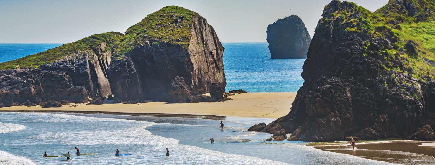 Playa de San Martín, una gran playa que con marea alta se convierte en varias calas. En primer plano se ve la orilla del mar con surfistas en el agua. Una isla y el acantilado de la costa se acercan estrechando la franja del arenal, para abrirse más allá. Al fondo en el mar, emerge otro islote de gran tamaño.