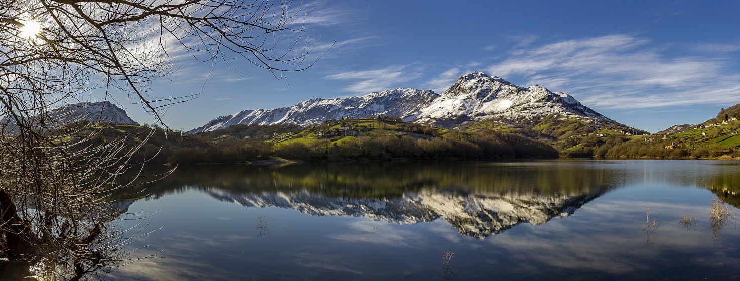 Fotografia da barragem de Alfilorios, com as suas águas a refletir como um espelho a montanha coberta de neve ao fundo. Em primeiro plano, uma árvore na margem esquerda da barragem. Outras montanhas mais baixas de ambos os lados da imagem, de cor verde e azulada, também se reflectem na água. Céu azul com algumas nuvens brancas.