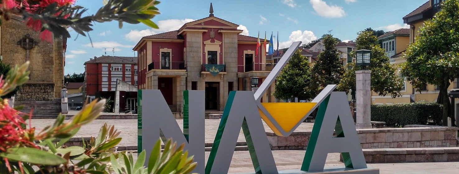 Fotografia di una piazza di Nava, in primo piano una scultura con le lettere NAVA, situata in una piazza pedonale. Sul lato sinistro alcuni fiori sembrano voler toccare le lettere.  Dietro c'è l'edificio del Municipio di colore marroncino, con le bandiere su un balcone, sulla destra alberi che chiudono la piazza e alcune case che si affacciano da dietro.
