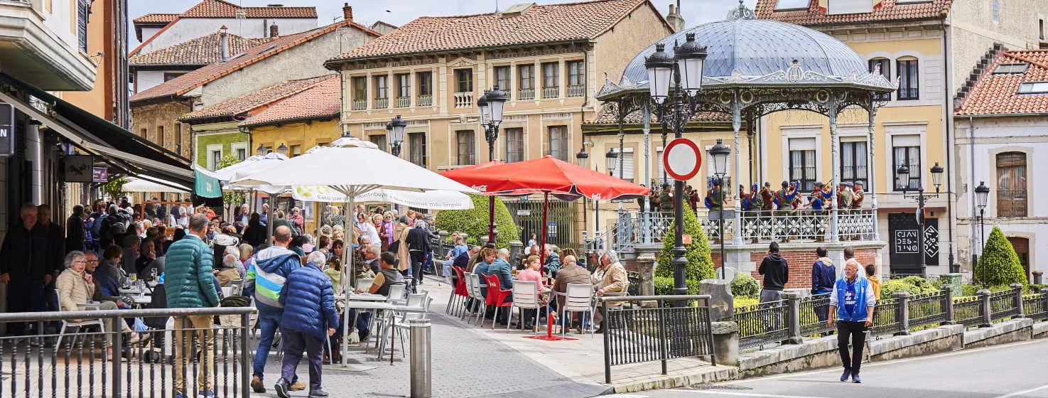 Picture of the square of Noreña celebrating one of its most famous festivals, the Picadillo and Sabadiego. In the centre is a bandstand with a military band, to the left are bars with terraces and colourful awnings and many people eating and drinking. In the background, brown, green and yellow buildings close the picture.