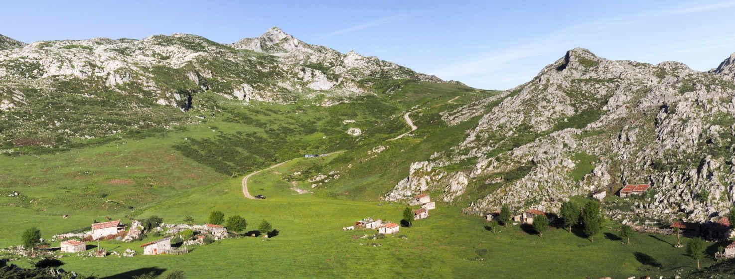 Bild der Majada de Balbín in Onís, einer grünen Wiese mit Hüttengruppen links und rechts des Bildes. In der Mitte führt ein Weg zum Pass, der zwei weitere Berggebiete mit grauem Kalkstein und grüner Macchia voneinander trennt.