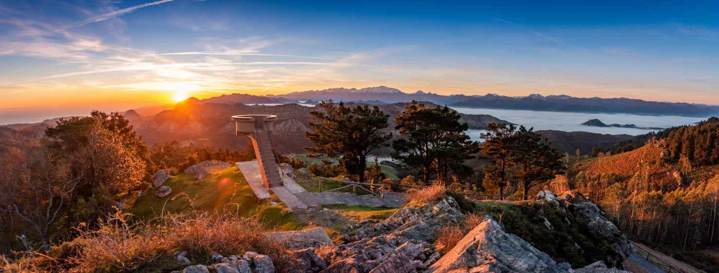 Amanecer en el Mirador del Fitu, el sol sale por el horizonte con sus rayos de luz roja rozando el horizonte, donde se ven los Picos de Europa con nieve en sus cumbres. A la izquierda al fondo, el mar. A la derecha un mar de nubes tapan el valle. En el centro, el mirador de Fitu y árboles alrededor.