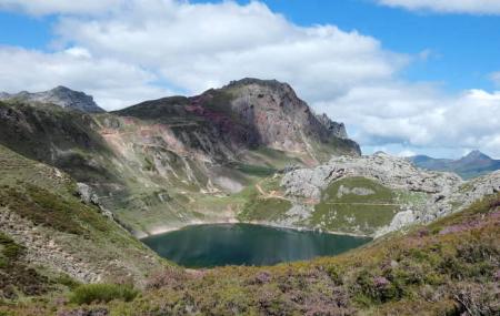 El Lago del Valle ocupa una cubeta de alta montaña rodeada de laderas rocosas y matorral, con senderos visibles en las pendientes. Al fondo se alza un macizo escarpado y el cielo está parcialmente nublado con luz diurna.