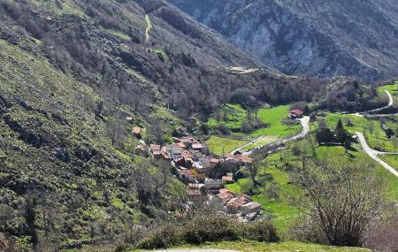 Vista de un pequeño pueblo en el fondo del valle, casas apiñadas con tejados rojos, una carretera llega al pueblo. A la izquierda una ladera con gran desnivel sube hacia las montañas más altas. Terreno de prados verdes, árboles otoñales y roca caliza. Los verdes intenesos se combinan con los marrones de los árboles y grises de la roca.