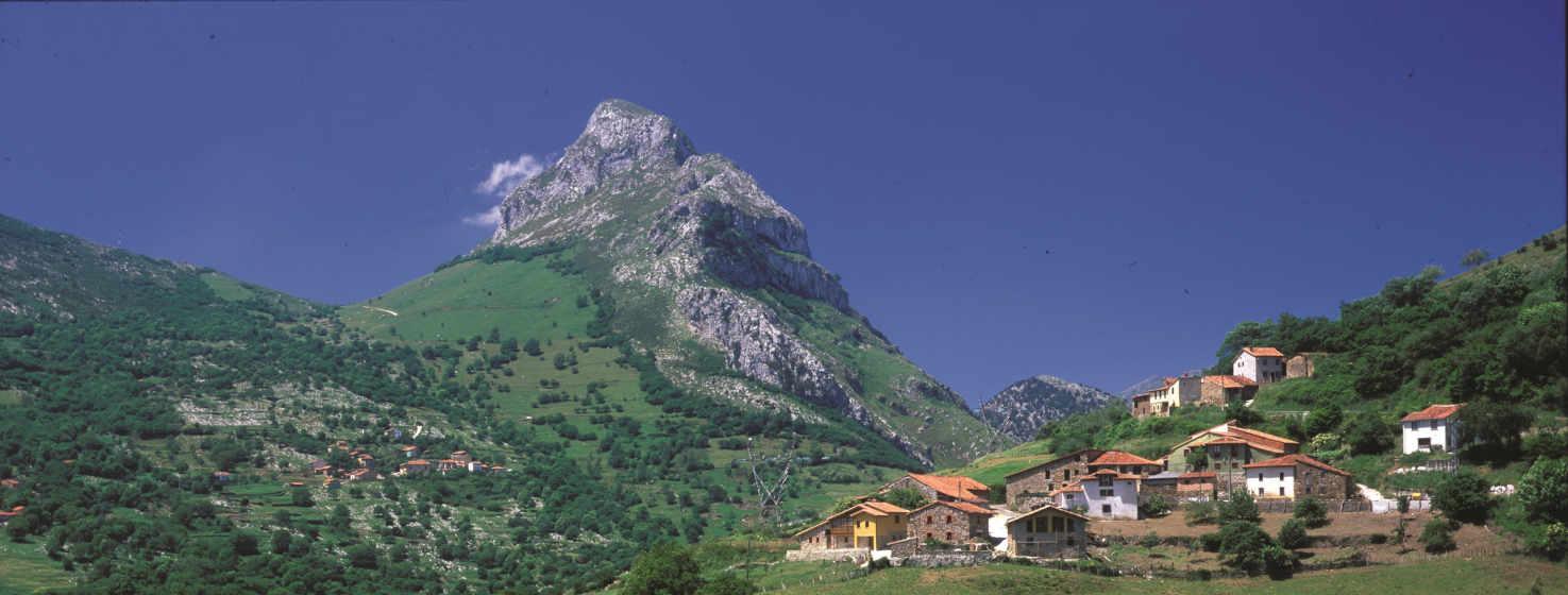Cenário de montanha, à direita da imagem uma pequena aldeia empoleirada na encosta da montanha, apenas a tocar o céu. No centro, ao fundo, ergue-se a Pica Peñamellera, uma imagem bem conhecida na zona, de rocha cinzenta e prados verdes. O azul intenso do céu contrasta com o resto das cores.