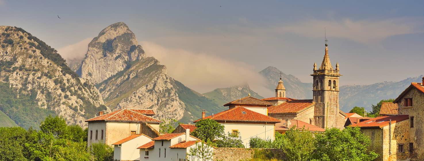 View of Alles, capital of Peñamellera Alta, a small mountain village with white houses with red roofs, in which the church with a tower stands out. It is surrounded by forests and mountains, which combine the grey or silver colours of the limestone typical of this area with the green of its meadows and the blue of the sky.