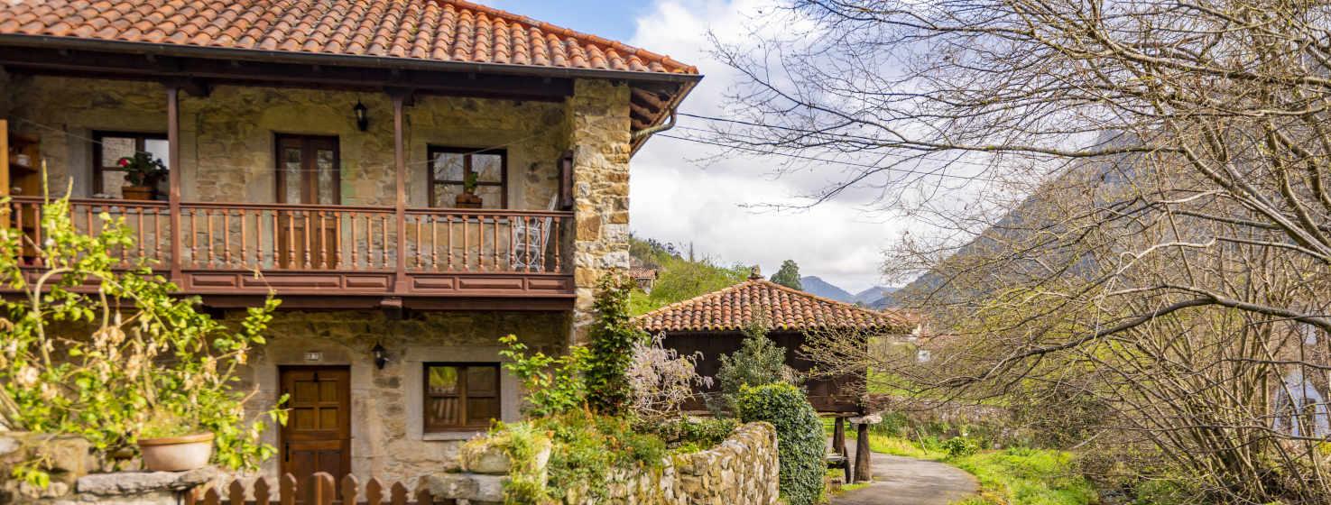 Image of a stone and wood house with an antojana and stone wall around it. It is on a road with a stream on the right. Behind the house a very well preserved granary. Dry trees in the foreground on the right and mountains behind.