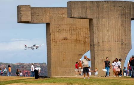 Avión ligero en vuelo a baja altura frente al Elogio del Horizonte, en Gijón, con varias personas paseando y observando en la zona verde del monumento bajo cielo parcialmente nublado.