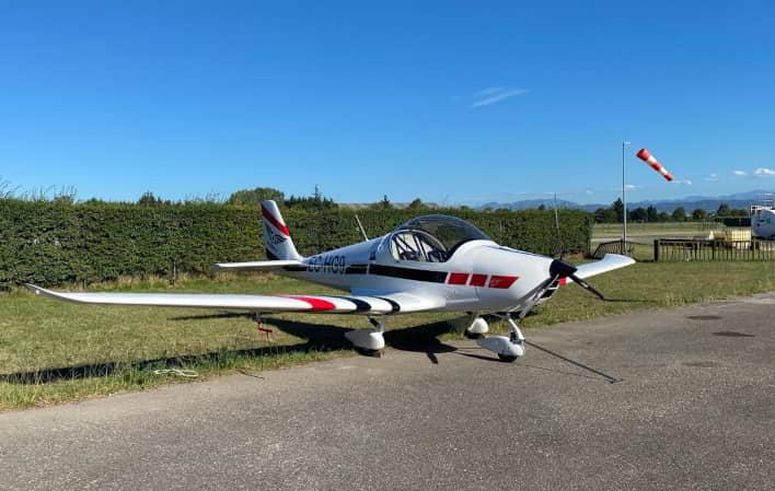 Avión ligero de dos plazas estacionado en una pista de aviación, con una manga de viento al fondo y cielo despejado en un entorno de pradera y seto bajo. Asturfly