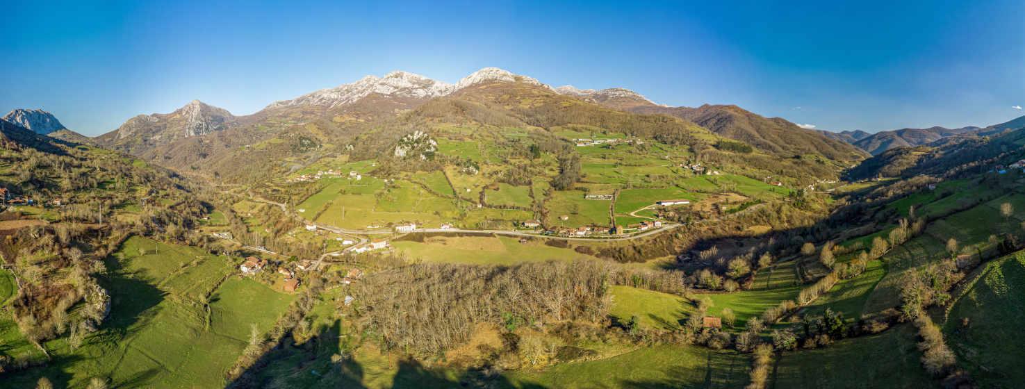 Bild des Tals von Quirós, weit mit Wiesen in verschiedenen Grüntönen, im Hintergrund Bergketten mit Büschen und etwas Schnee auf ihren Gipfeln. Der Himmel ist klar, ganz blau. In der Talsohle ist eine Straße zu sehen, die das Tal durchquert, und ein kleines Dorf auf beiden Seiten der Straße.