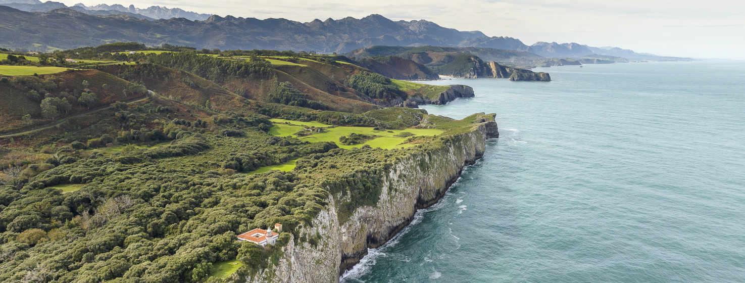 Vista de la costa de Ribadedeva con el faro de San Emeterio al borde del acantilado.  Impresionantes paredes de roca caliza caen al mar. Encima praderías verdes, con árboles a lo largo de la costa. Al fondo se pueden ver cadenas montañosas, en tonos más oscuros, y detrás se ven los Picos de Europa con sus colores más grises de la roca.