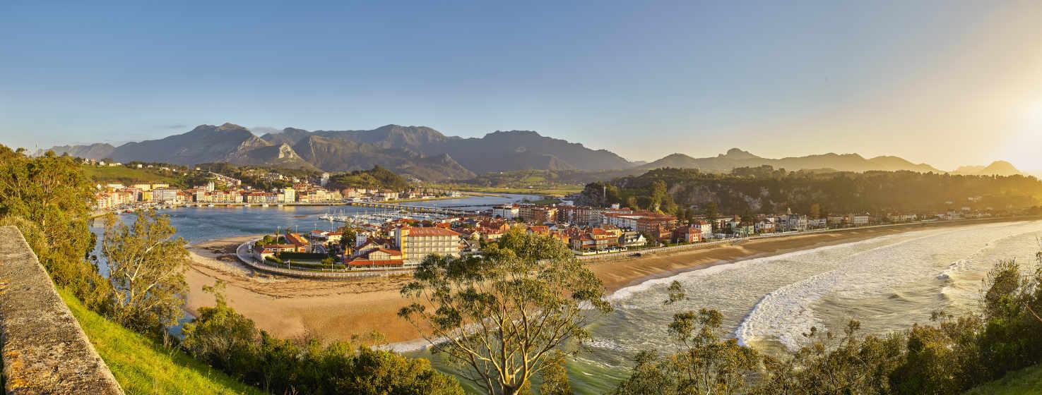 Vista panorâmica de Ribadesella. Vê-se a praia com o seu passeio marítimo e a zona de moradias e hotéis. O estuário que se junta ao mar para formar outra pequena zona de areia. Na parte de trás do estuário, à direita, encontra-se o porto desportivo e, à esquerda, o porto de pesca e a cidade. Por detrás das montanhas que dominam a imagem, tocando o céu azul.
