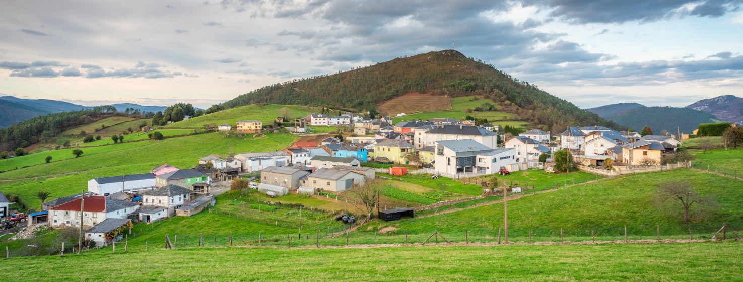 Fotografia geral da aldeia de Villayón. Situada numa colina verde, as suas casas são brancas com telhados de ardósia cinzenta. Atrás da aldeia ergue-se uma pequena montanha coberta de árvores não muito altas em tons verdes e castanhos mais escuros. Ao fundo, mais cadeias de montanhas de cores mais escuras e azuladas fecham o quadro. O céu com nuvens e céu limpo.