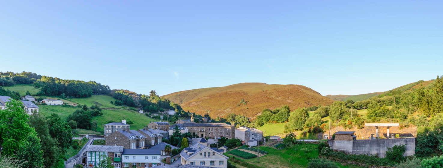 Panoramic view of Vilanova, capital of the municipality of Villanueva de Oscos, a peaceful rural village at the foot of rolling hills bathed in warm sunlight. Stone houses and slate roofs blend harmoniously into the landscape, surrounded by green meadows and lush forests.