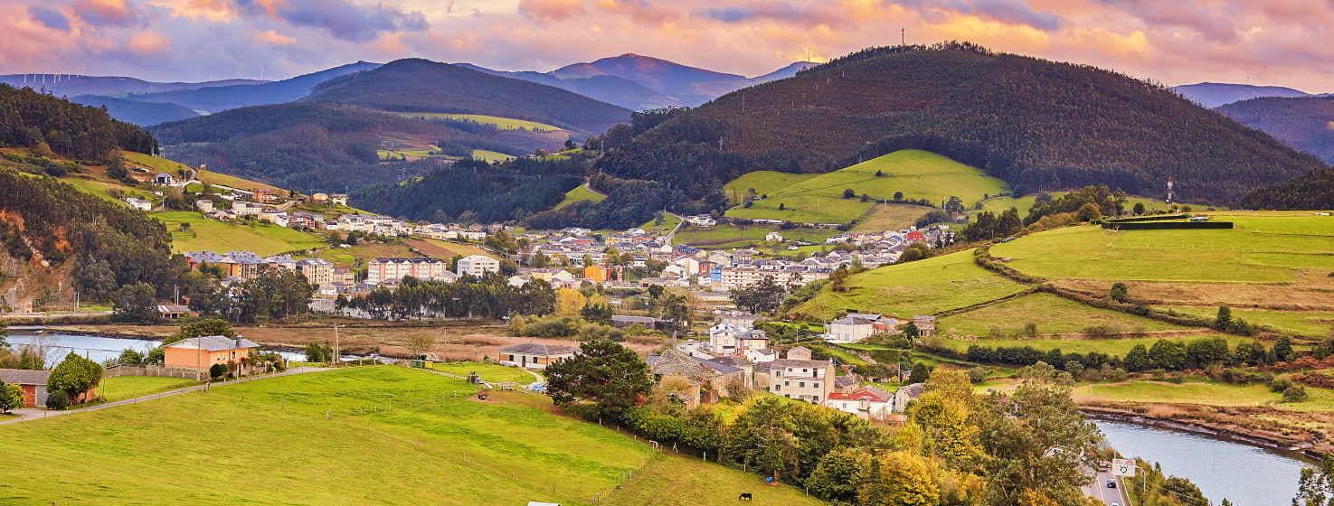 General view of Vegadeo. Some green meadows in the foreground with some trees and houses. The estuary runs in front of the village, which expands into the valley. Around it rise hills covered with trees and behind it higher mountains in shades of blue and purple. The dark clouds in the sky give the picture a wintry look.