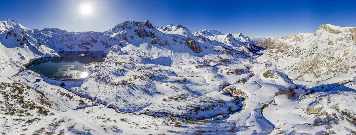 Vista panorámica de un circo glacial cubierto de nieve, con su gran lago rodeado de altas montañas que dejan ver sus rocas entre la nieve. Un camino sube valle arriba hasta el lago, algunos árboles y arbustos se ven salpicados por todo el paisaje. Al fondo del valle se aprecia el pueblo de Lago del Valle. El sol reluciente reina en la imagen.
