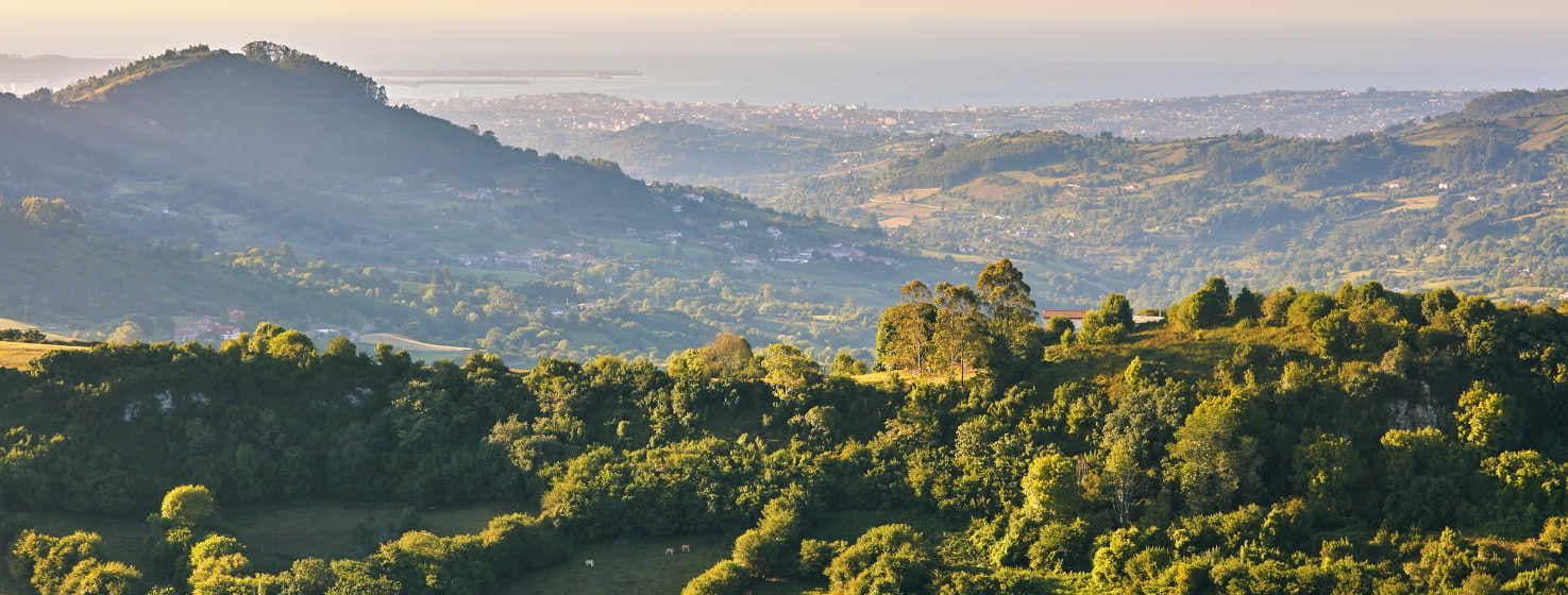 View from the summit of Peña Careses. In the foreground an area with farms and trees and a house with a red roof. In the background you can see the coast of Gijón and the sea. In the middle the valleys of the area with small nuclei of houses and gentle hills.