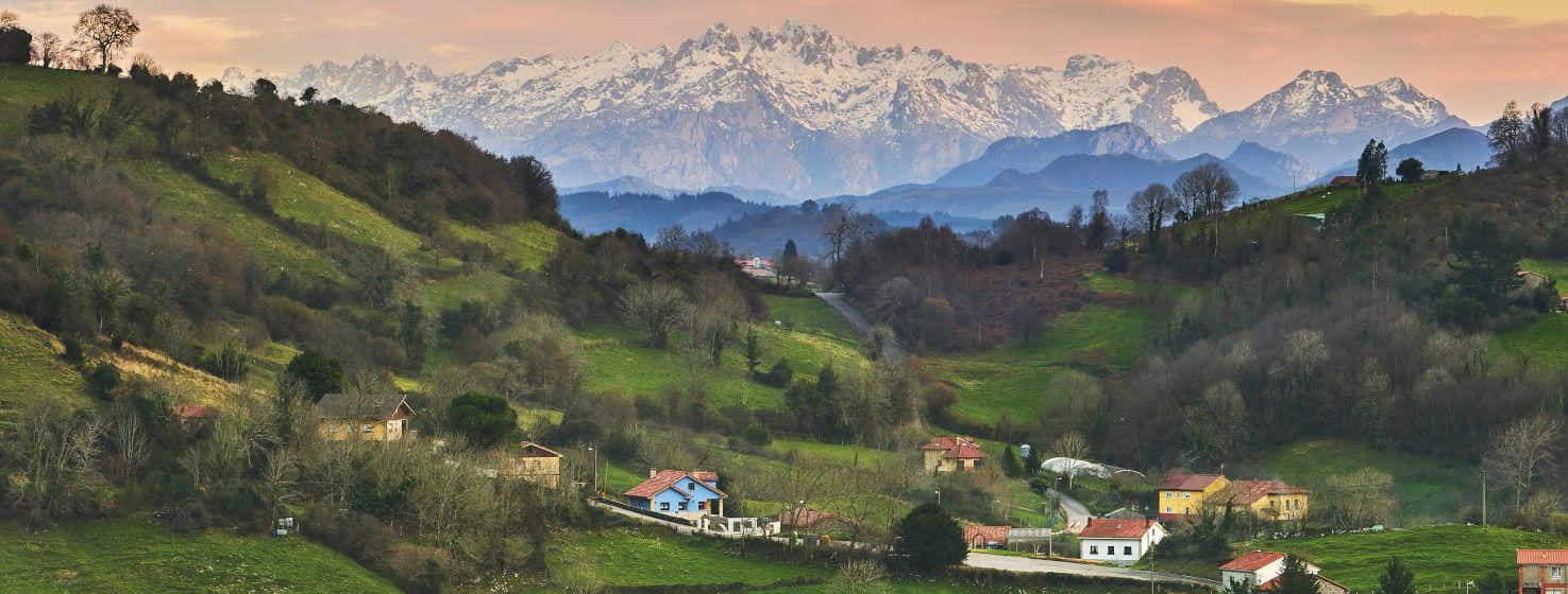 View of a valley of Sariego, full of green farms with some trees, you can see some houses of different colours, blue, white, yellow and red roofs.  In the background there are several mountain ranges in shades of blue, ending with a view of the snow-capped Picos de Europa, brushing the sky which has an orangey sunset colour with some grey clouds.