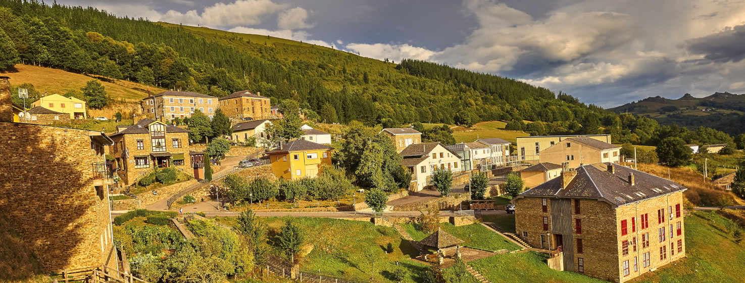 Vista di Santalla, capoluogo di Santa Eulalia de Oscos, un pittoresco villaggio asturiano. Le case dai colori vivaci si aggrappano alle pendici delle colline, creando un mosaico di facciate che contrastano con la vegetazione lussureggiante. Sullo sfondo si staglia un paesaggio montuoso, mentre un cielo azzurro con nuvole bianche aggiunge luminosità alla scena.